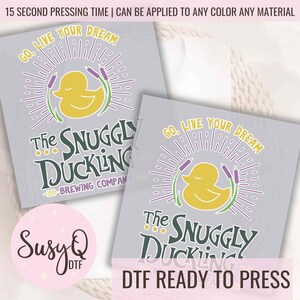 May include: A digital transfer design featuring a yellow rubber ducky with purple reeds and a green sunburst. The text "Go. Live Your Dream" and "The Snuggly Duckling Brewing Company" is displayed in a green font. The design is ready to be pressed onto a t-shirt or other fabric.