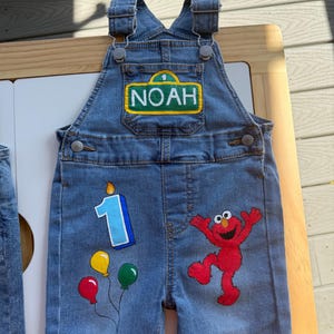 May include: Blue denim overalls featuring a rectangular patch on the chest with the name NOAH and the number 1. The front of the legs displays a graphic of the character Elmo, a blue number 1 candle, and three colorful balloons in red, yellow, and green.