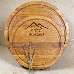 Könnte beinhalten: Drei runde Holzservierbretter mit einer gefrästen Nut am Rand. Die Bretter sind hellbraun mit Holzmaserung. Das mittlere Brett trägt den Text "Papa's Brotzeit" und eine Berg-Grafik. Ein Lavendelstrauß liegt davor.