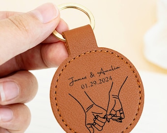 Könnte beinhalten: Ein brauner Leder-Schlüsselanhänger mit goldenem Ring. Der runde Anhänger zeigt eine Strichzeichnung von zwei Händen, die sich halten, mit den Namen "James & Amelia" und dem Datum "01.29.2024" aufgedruckt.