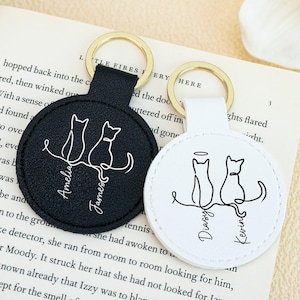 May include: Two personalized cat keychains. One is black with white text and a gold keyring, the other is white with black text and a gold keyring. Each keychain features a line drawing of two cats.