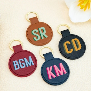 Peut inclure: Quatre porte-clés personnalisés en cuir marron, bordeaux, marine et noir. Chaque porte-clés est orné d'un anneau doré et gaufré d'initiales aux couleurs contrastées : vert, bleu clair, orange et rose. Les porte-clés sont ronds et ont un bord cousu.
