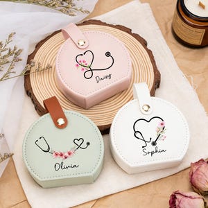 May include: Three round, pastel-colored cases with a stethoscope design and floral accents. Each case has a different name: Daisy, Olivia, and Sophia. The cases have a handle and are on a wooden slice.