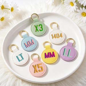 May include: Seven pastel-coloured round keyrings with gold-coloured rings. Each keyring features a different combination of letters and numbers in contrasting colours, including "X3", "MM", "1404", and "LT".