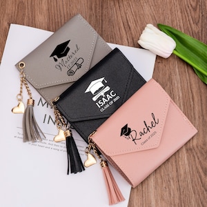 May include: Four small graduation wallets in gray, black, and pink. Each wallet has a tassel and heart charm. The wallets are personalized with names, graduation caps, and the text "Mastered IT 2025", "ISAAC CLASS OF 2023", and "Rachel Class of 2025".