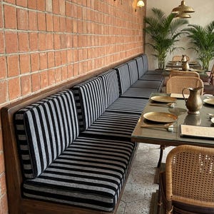 May include: A restaurant interior featuring a long wooden bench with black and gray striped cushions. The bench is set against a brick wall, with tables and chairs visible in the background. Brass light fixtures hang from the ceiling.