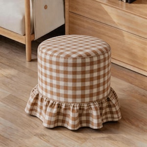 Op de afbeelding: Een ronde poef bekleed met een beige en witte gingham ruitjesstof. De cilindrische zitting is voorzien van een geplooide, gerimpelde rok rond de basis, waardoor een zachte, geplooide rand ontstaat die uitsteekt vanaf het hoofdmeubel.