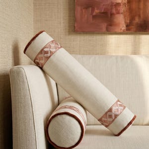 May include: Two cylindrical decorative pillows on a cream-colored sofa. The pillows are off-white with a geometric pattern in a light pink hue and brown trim. The pillows are leaning against the sofa's armrest.