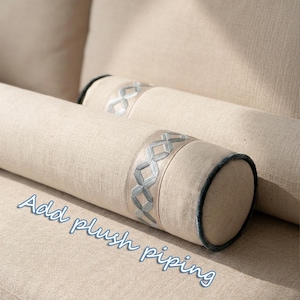 May include: Two beige bolster pillows with decorative silver geometric trim and dark blue piping. The pillows are cylindrical and rest on a matching beige surface. The text "Add plush piping" is written in light blue cursive.