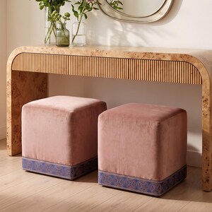 May include: Two square ottomans upholstered in dusty pink velvet fabric. Each ottoman features a wide, decorative band at the base in a deep blue color with a repeating circular geometric pattern in a lighter blue and gold tone.