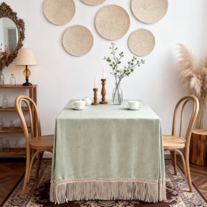 Könnte beinhalten: Esszimmer-Szene mit einer salbeigrünen Tischdecke mit Fransen, gedeckt mit weißen Teetassen und Kerzen. Gewebte Wanddekoration und Holzstühle vervollständigen die gemütliche, neutral gehaltene Ästhetik.