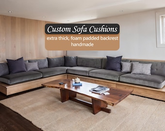 May include: A large corner sofa with grey cushions and scatter cushions. The sofa is built into a wooden base. A wooden coffee table sits on a beige rug in front of the sofa. Text reads "Custom Sofa Cushions extra thick, foam padded backrest handmade."