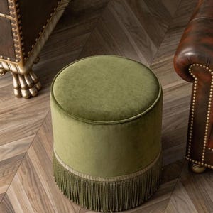 May include: A round, olive green velvet ottoman featuring a smooth, padded top and a decorative fringe trim around the base. The cylindrical stool is upholstered in a soft, solid-coloured fabric and stands as a standalone piece of furniture.