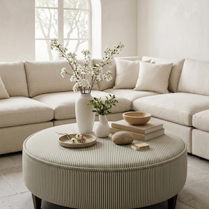 Puede incluir: Un otomán redondo tapizado en tela de rayas finas beige y blanca con patas de madera oscura. Encima hay una bandeja de cerámica beige con flores pequeñas, dos jarrones blancos con flores blancas y hojas verdes, un cuenco de madera, libros y una piedra.