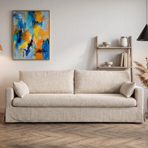 May include: A beige fabric sofa with two large back cushions and two small side pillows. The sofa features a skirted base and is positioned in a room with a floor lamp, a wooden shelving unit, a wall painting, and a vase with dried plants.
