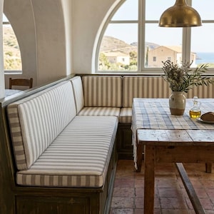 May include: A built-in corner bench with striped cushions and a wooden base. A wooden table with a checkered tablecloth is set near a large arched window. A brass pendant light hangs above the table.