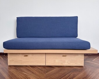 May include: A contemporary sofa with a navy blue cushion and backrest. The base is made of light-coloured wood and has two drawers with small, gold-coloured handles. The sofa is positioned on a wooden floor.