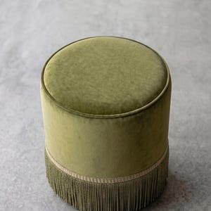 K&ouml;nnte beinhalten: Ein runder Hocker, der mit olivgr&uuml;nem Samtstoff bezogen ist. Der zylindrische Sitz verf&uuml;gt &uuml;ber eine flache Oberseite und eine Basis, die mit einem breiten Band aus passenden olivgr&uuml;nen Fransen verziert ist, die bis zum Boden reichen.
