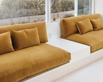 May include: Two mustard-coloured built-in sofas with matching cushions. The sofas are positioned against a large window, creating a comfortable seating area. The design is minimalist and modern, with clean lines and a focus on comfort.