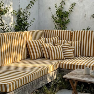 May include: Outdoor sectional sofa with mustard yellow and white striped cushions and pillows. The sofa is made of wood and is surrounded by greenery and a white wall. A small wooden table is in front of the sofa.