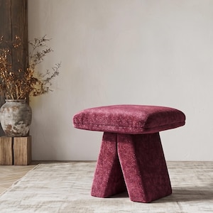 Puede incluir: Un taburete cuadrado tapizado en una tela texturizada de color magenta intenso. El taburete cuenta con un asiento grueso y acolchado sostenido por una base sólida en forma de V que se ensancha hacia el suelo, creando un mueble cohesivo.