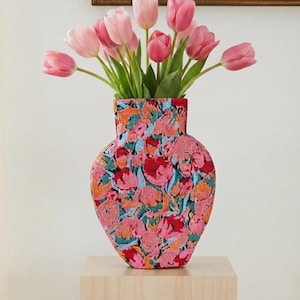 May include: A vase with a flat, heart-shaped silhouette covered in a textured, multi-coloured floral pattern of pink, orange, and teal threads. The vase is filled with a bouquet of fresh pink tulips with green stems and leaves.