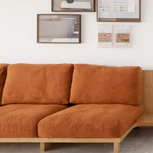 May include: A contemporary sofa featuring a wooden frame and burnt orange cushions. The sofa has a minimalist design, with clean lines and a built-in side table. The cushions appear to be made of a textured fabric. The sofa is against a white wall with framed artwork.