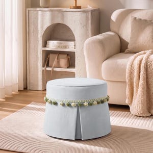 May include: A round, light blue upholstered ottoman featuring a pleated skirt design and a decorative trim of small, sage green pom-poms encircling the upper edge. The ottoman has a flat, circular top and a structured, cylindrical base.