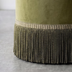 K&ouml;nnte beinhalten: Ein zylindrischer Hocker, der mit olivgr&uuml;nem Samtstoff bezogen ist. Die Basis des Hockers ist mit einer breiten, gewebten Borte in Beige und Braun verziert, an der eine lange, dichte Franse aus passenden olivgr&uuml;nen gedrehten Kordeln h&auml;ngt.