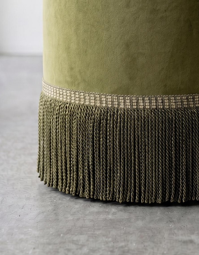 K&ouml;nnte beinhalten: Ein zylindrischer Hocker, der mit olivgr&uuml;nem Samtstoff bezogen ist. Die Basis des Hockers ist mit einer breiten, gewebten Borte in Beige und Braun verziert, an der eine lange, dichte Franse aus passenden olivgr&uuml;nen gedrehten Kordeln h&auml;ngt.