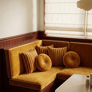May include: A mustard yellow velvet corner booth with a dark wood base, featuring two square striped throw pillows in shades of red and gold, and two round tufted mustard yellow velvet cushions, positioned next to a light wood table.