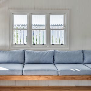 May include: A light blue sectional sofa with multiple cushions sits on a wooden base. The sofa is positioned beneath a triple-pane window with white trim and a white picket fence design outside.