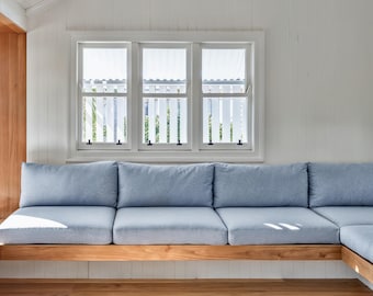 May include: A light blue sectional sofa with multiple cushions sits on a wooden base. The sofa is positioned beneath a triple-pane window with white trim and a white picket fence design outside.