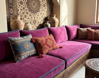 May include: A long, low-profile sofa upholstered in deep magenta velvet with teal piping, featuring a carved wooden base. It is adorned with several decorative throw pillows in blue, red, and patterned fabrics, set against a wall with a large tapestry.