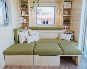 May include: Interior view of a contemporary living area featuring a green sectional sofa and integrated shelving. The sofa is decorated with white and patterned cushions. Shelves showcase ornaments and plants, creating a comfortable and stylish ambience.
