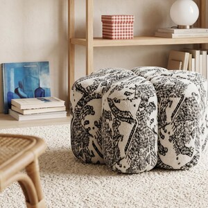 May include: A low, four-lobed ottoman featuring a cream-colored fabric base with a dense, black ink-style illustrative pattern depicting various abstract shapes and figures. The piece has a rounded, segmented silhouette with a soft, textured surface.