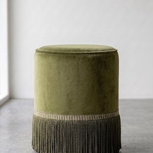 K&ouml;nnte beinhalten: Ein zylindrischer Hocker, bezogen mit olivgr&uuml;nem Samtstoff. Der Sockel des Hockers ist mit einem dekorativen gewebten Band und langen, passenden olivgr&uuml;nen Fransen versehen, die bis zum Boden reichen und den unteren Teil des M&ouml;belst&uuml;cks bedecken.