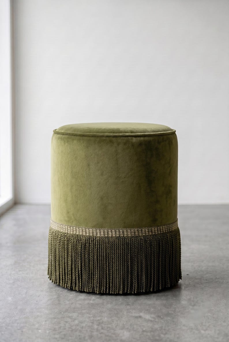 K&ouml;nnte beinhalten: Ein zylindrischer Hocker, bezogen mit olivgr&uuml;nem Samtstoff. Der Sockel des Hockers ist mit einem dekorativen gewebten Band und langen, passenden olivgr&uuml;nen Fransen versehen, die bis zum Boden reichen und den unteren Teil des M&ouml;belst&uuml;cks bedecken.