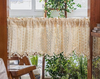 May include: A cream-colored crocheted valance with a scalloped bottom and tassel fringe hangs in a window. The valance is made of delicate lace and has a floral pattern.