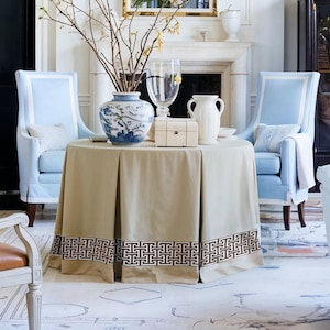 May include: A living room scene featuring a round table draped with a tan tablecloth with a decorative border. Two light blue armchairs flank the table, and a vase with branches sits atop it. A fireplace and other furniture complete the elegant setting.