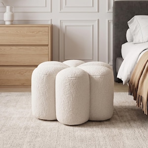 May include: A cream-colored, flower-shaped ottoman upholstered in a textured, fuzzy fabric. The piece features six rounded, cylindrical segments arranged in a circular pattern, creating a soft, plush appearance with a low-profile design.