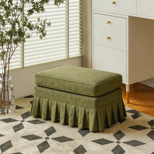 May include: A rectangular olive green ottoman featuring a thick, cushioned top and a pleated skirt base. The fabric has a visible woven texture, and the piece is designed with clean, straight lines, providing a structured appearance for seating.