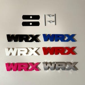 May include: A collection of WRX emblems in various colors, including black, blue, white, red, pink, and silver. The image also shows mounting hardware and black mounting brackets. The emblems are arranged on a white surface.