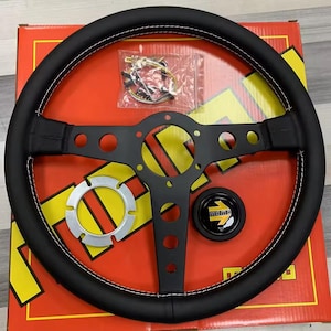 Könnte beinhalten: Schwarzes Lederlenkrad mit weißen Nähten und einem Momo-Logo. Das Lenkrad hat eine schwarze Mittelnabe mit sechs Speichen und eine silberne Hupe.