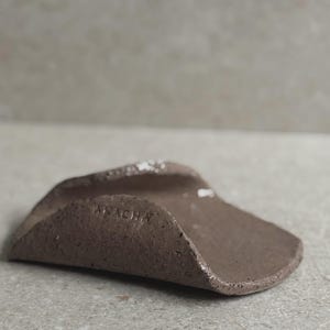 Könnte beinhalten: Eine kleine, handgefertigte, braune Keramikschale mit einem gebogenen, erhöhten Rand. Die Schale hat eine strukturierte Oberfläche und das Wort "ALACHA" ist auf der Innenseite eingeprägt. Die Schale ist für kleine Gegenstände konzipiert.