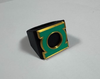 May include: A black ring featuring a square face with a green square inlay and a gold-colored circular emblem in the center. The design includes a raised gold-colored border around the green square and the central circular cutout.