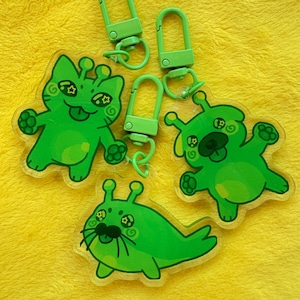 May include: Green acrylic keychains featuring cartoon alien characters with star and swirl details. Each keychain has a matching green clasp. The keychains are on a yellow background.