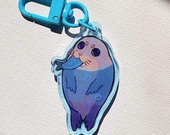 May include: A clear acrylic keychain featuring a cartoon seal with a light purple head and a blue body, holding a small blue fish in its mouth. The keychain is attached to a bright blue plastic lobster clasp hardware for easy attachment.