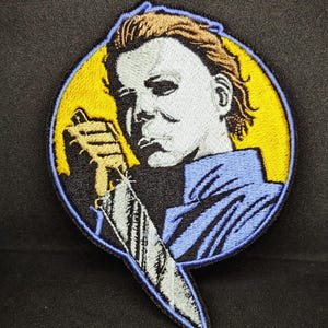 May include: Embroidered patch featuring a character from the horror movie Halloween. The character is depicted with a white mask, brown hair, and a blue shirt. He is holding a knife with a silver blade. The background is yellow.