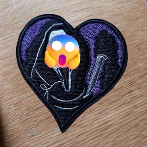 May include: Heart-shaped embroidered patch with a black Grim Reaper design holding a scythe, set against a purple and black background. The patch has a stitched border, suitable for clothing or accessories.